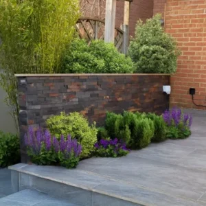 A modern garden with Jak Multi Stone Cladding on the raised planter, green shrubs, and purple flowers beside gray tiled steps. A wooden pergola and a brick wall add style to the background.