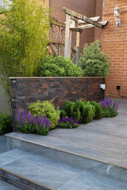 A modern garden with Jak Multi Stone Cladding on the raised planter, green shrubs, and purple flowers beside gray tiled steps. A wooden pergola and a brick wall add style to the background.
