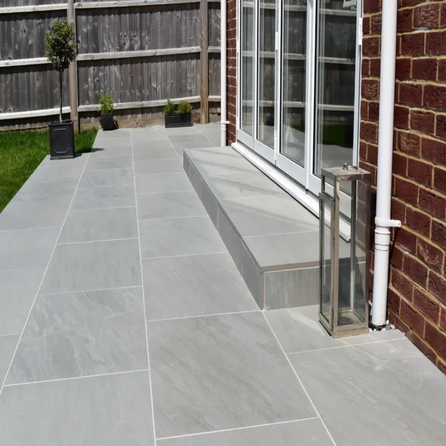 A contemporary patio showcases Kandla Grey Outdoor Porcelain Paving Tiles in a 600mm x 900mm size, forming a sleek expanse. It features a single step leading to stylish glass patio doors and is complemented by an adjoining brick wall. The area is enclosed by a wooden fence and adorned with several potted plants. A lantern-style light fixture is elegantly placed on the tiles.