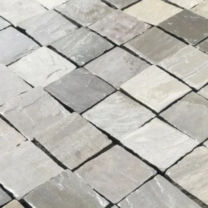 Close-up of a paved surface composed of square Kandla Grey Indian Sandstone Cobble Setts, 100mm x 100mm x 40/60mm. The setts feature varying shades of light grey and beige, creating a textured and uniform pattern across the image.