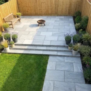 Modern garden with Kandla Grey Indian Sandstone Paving slabs (18mm Calibrated, 600mm x 900mm), a green lawn, potted plants, wooden bench, fire pit, and privacy provided by wooden fencing.