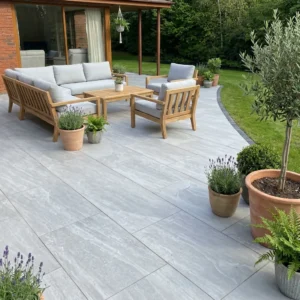 A modern patio features Kandla Grey Outdoor Porcelain Paving Tiles (600mm x 900mm x 20mm), wooden outdoor furniture with gray cushions, potted plants, green grass, trees, and a glass sliding door into a brick house.