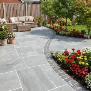 A landscaped backyard patio features Kandla Grey Indian Sandstone (18mm Calibrated Mixed Size) tiles, wicker outdoor furniture with beige cushions, potted plants, and curved flower beds of colorful flowers along a wooden fence.