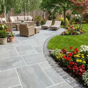 A landscaped backyard with a Kandla Grey Indian Sandstone patio, wicker furniture, lounge chairs, potted plants, colorful flower beds bordered by lawn and a wooden fence. Shade and greenery are provided by trees.