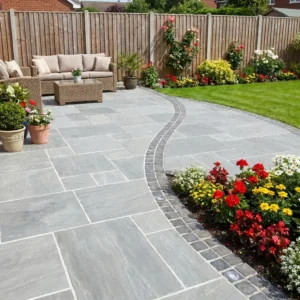 A neatly landscaped backyard features a stone patio made with Kandla Grey Indian Sandstone - 18mm Calibrated Mixed Size, outdoor wicker furniture, potted plants, colorful flower beds, green lawn, and a wooden fence in the background.