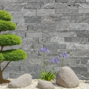 Monsoon Black Stone Cladding creates a striking backdrop for a minimalist garden featuring trimmed green shrubs, blooming purple flowers, river rocks, and white gravel.