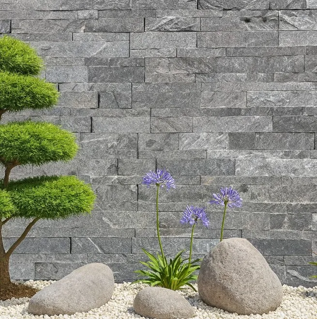 Monsoon Black Stone Cladding creates a striking backdrop for a minimalist garden featuring trimmed green shrubs, blooming purple flowers, river rocks, and white gravel.
