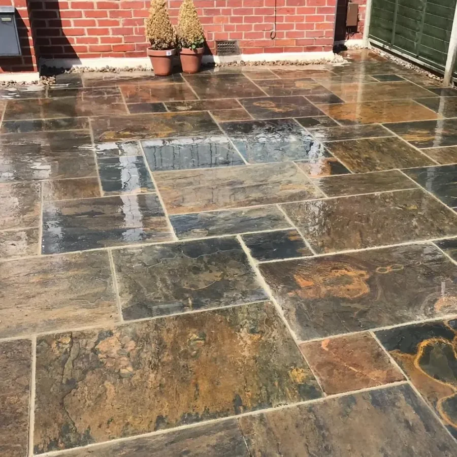 A damp patio showing Copper Slate 20mm Calibrated Mixed Size tiles in earthy hues of brown, gray, and orange. Two potted plants with cone-shaped shrubs are positioned near a brick wall in the background, and the surface beautifully reflects the sky.