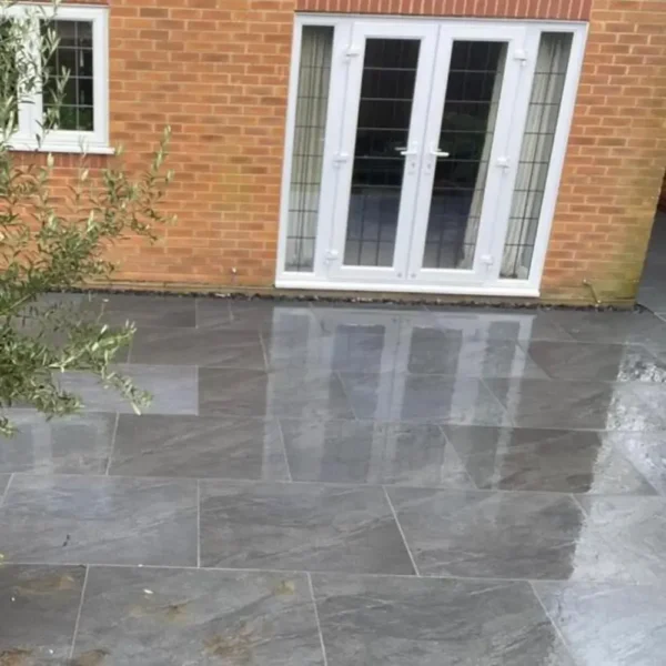 A damp outdoor area with County Black Anthracite Outdoor Porcelain Paving Tiles in dark gray is situated in front of a brick building. There are white-framed double doors and a window, while an olive tree with lush green leaves is partially visible on the left side.