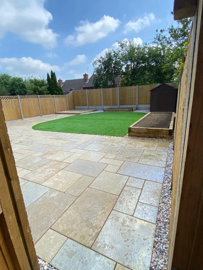 A newly landscaped backyard features a circular patch of green artificial grass in the center, surrounded by stone paving. A wooden fence encloses the area, and a small garden shed is visible on the right. The sky is partly cloudy.