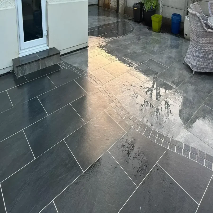 A modern patio showcasing large Brazilian Black Outdoor Porcelain Paving Tiles, measuring 600mm x 900mm x 20mm, with a polished finish. These dark tiles are beautifully bordered by smaller, lighter tiles. A wicker chair rests against a pristine white wall, and several vibrant pots filled with plants are arranged near a partition.