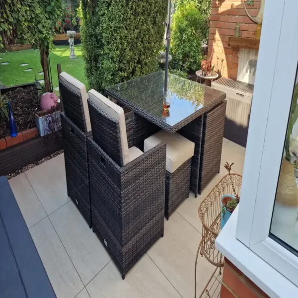 Scene depicting a patio with Latte Outdoor Porcelain Paving Tiles (600mm x 900mm x 20mm), complete with a wicker dining set featuring a glass-topped table and cushioned chairs, all shaded by a large umbrella. The area is enveloped by greenery, along with a lush garden in the background, as a drink sits on the table to add a relaxed touch to this serene setting.
