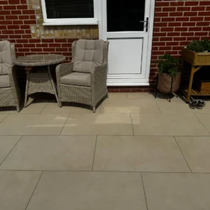A patio with cushioned wicker chairs and a round table sits on Aosta Crema Outdoor Porcelain Paving Tiles (600x900x20mm) in front of a brick wall with a white door, alongside a wooden planter, potted plant, and shoes.