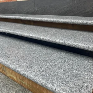 Close-up of Silver Grey Granite Bullnose Steps 900x350x35mm—three outdoor steps with a rough, speckled gray finish, bordered by wooden supports and leading up to a brick wall and dark stone tiles.