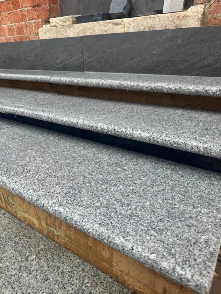 Close-up of Silver Grey Granite Bullnose Steps 900x350x35mm—three outdoor steps with a rough, speckled gray finish, bordered by wooden supports and leading up to a brick wall and dark stone tiles.
