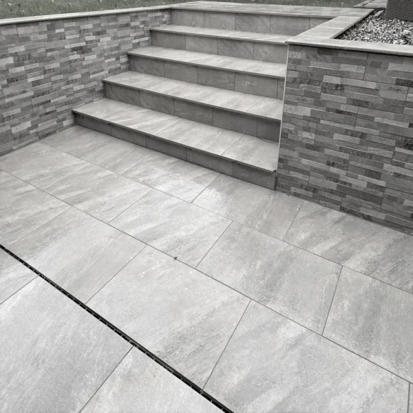 A set of Quartz Grey Outdoor Porcelain Paving Tiles, sized at 600mm x 900mm x 20mm, forms the steps leading up from a matching patio. The surrounding walls feature textured stone tiles that provide contrast to the smooth, large floor tiles. A small planted area is visible at the top right.