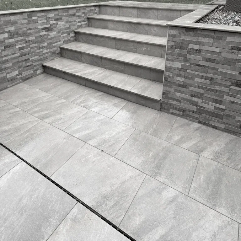 A set of Quartz Grey Outdoor Porcelain Paving Tiles, sized at 600mm x 900mm x 20mm, forms the steps leading up from a matching patio. The surrounding walls feature textured stone tiles that provide contrast to the smooth, large floor tiles. A small planted area is visible at the top right.