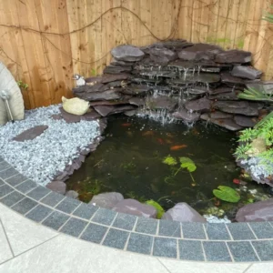 A small backyard pond with a cascading waterfall surrounded by shale stones and Blue/Black Granite Sawn Setts measuring 100mm x 100mm x 40mm. Decorative elements include a leaf-shaped sculpture, pebbles, and leafy plants. The pond features lily pads and glimpses of fish swimming, with a wooden fence in the background.