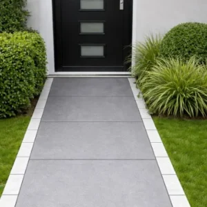 A modern front walkway paved with large gray tiles and bordered by Quartz White Outdoor Porcelain Cobble Setts (100x200x20mm), leading to a black door with four glass panels, is flanked by green bushes and grass on both sides.