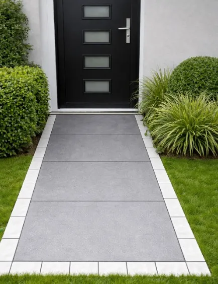 A modern front walkway paved with large gray tiles and bordered by Quartz White Outdoor Porcelain Cobble Setts (100x200x20mm), leading to a black door with four glass panels, is flanked by green bushes and grass on both sides.