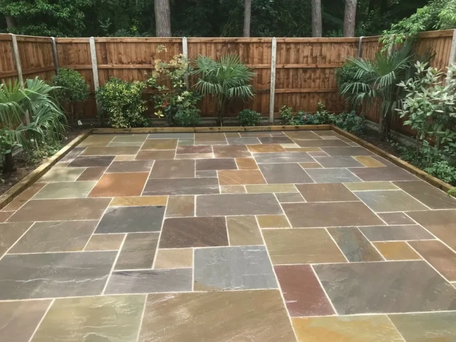 A neatly arranged stone patio with rectangular and square tiles in earthy tones. The patio is bordered by a wooden fence and surrounded by various plants and small trees.