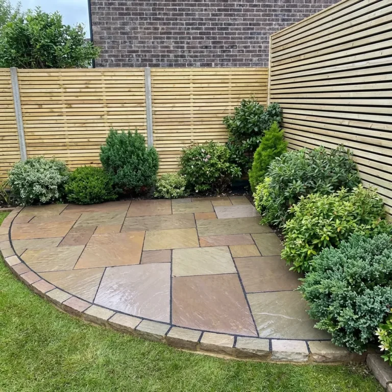 A small garden corner showcases a curved patio laid with Raj Blend Indian Sandstone Paving Slabs (riven, 18mm, mixed size), bordered by green shrubs and bushes, wooden slat fencing, a brick wall backdrop, and grass around the edge.