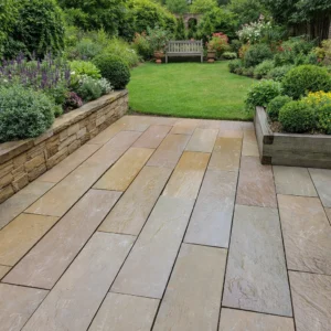 A patio made with Raj Green Indian Sandstone Paving Planks (riven, 900x200x22mm) leads to a lush garden of green grass, vibrant flowers, manicured bushes, a wooden bench, and leafy trees and plants.