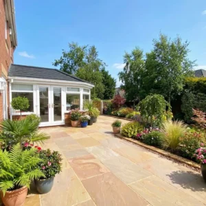 A sunny patio with potted plants and flowers features Raj Blend Indian Sandstone Paving Slabs (Riven, 18mm Calibrated, 600x900mm), leading to a lush garden with trees, shrubs, and vibrant flowerbeds under a clear blue sky.