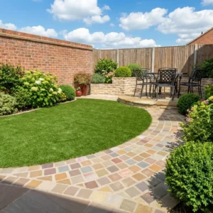 A neatly landscaped backyard features a small lawn, flower beds, and a raised patio with seating, plus a curved pathway made from Raj Green Cobble Setts 100mm x 100mm x 25-35mm, all bordered by brick and wooden fences under a blue sky.