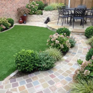 A landscaped backyard features a curved path of Raj green sandstone cobbles 100mm x 100mm, manicured shrubs, blooming flowers, artificial grass, a patio with a metal dining set, and a small black dog sitting near a raised flower bed.
