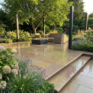 A patio featuring Raj Green Honed Sandstone Bullnose Steps (900mm x 350mm x 35mm), wicker outdoor furniture, a black coffee table, and blooming flower beds. Surrounded by trees and greenery, it creates a peaceful garden ambiance.