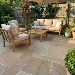 Outdoor raj green sandstone patio with light brown stone tiles, wooden furniture set with beige cushions, a coffee table, various potted plants, and a wooden fence with greenery in the background.