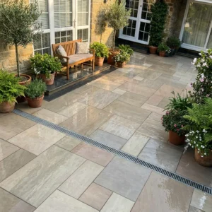 A tidy patio featuring Raj Green Outdoor Porcelain Paving Tiles in mixed sizes, potted plants, a wooden bench with cushions, large windows, and a tan brick wall framing the freshly cleaned area.