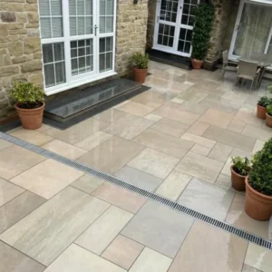 A tidy outdoor patio features Raj Green Outdoor Porcelain Paving Tiles in mixed sizes, potted plants along the edges, a central drainage grate, and glass doors opening into a stone-walled house.