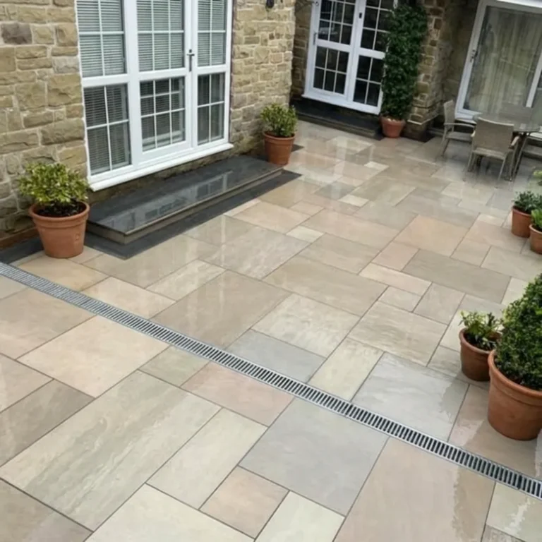 A tidy outdoor patio features Raj Green Outdoor Porcelain Paving Tiles in mixed sizes, potted plants along the edges, a central drainage grate, and glass doors opening into a stone-walled house.