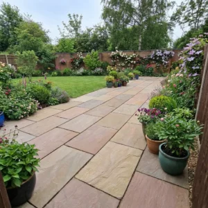 A stone patio made of Raj Green Indian Sandstone Paving Slabs (600x900mm, handcut, 22mm calibrated) is lined with potted plants and leads to a lush lawn bordered by vibrant flowers and shrubs near a wooden fence.