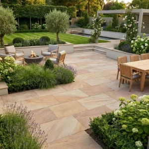 Beautiful landscaped backyard with stone patio, outdoor dining table, seating area around a fire pit, lush greenery, flowering plants, trimmed hedges, and a pergola in the background.