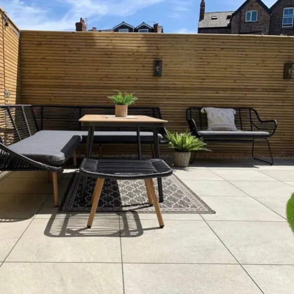 A contemporary outdoor patio framed by a wooden privacy fence showcases a gray cushioned bench, a black bench, and a small table adorned with a potted plant. A separate small plant sits on the ground beside the textured rug, all set atop HS Beige Outdoor Porcelain Paving Tiles in 600mm x 600mm x 20mm. Beyond the fence, brick houses are visible.