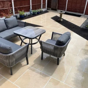 A patio with HS Beige Outdoor Porcelain Paving Tiles - 600mm x 600mm x 20mm showcases a seating area that includes a gray cushioned sofa, two armchairs, and a square table. Potted plants enhance the background, while a round birdbath is positioned near the fence. The area is enclosed by red wooden fencing.