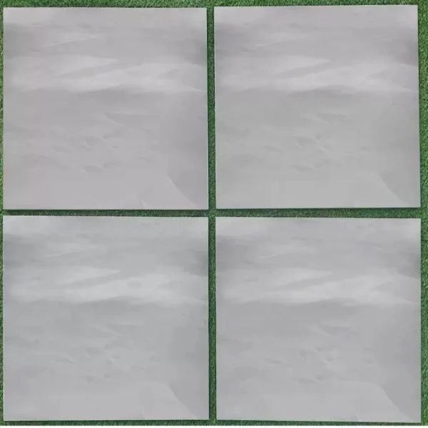 Four Brazilian Grey Slate Outdoor Porcelain Paving Tiles, each measuring 600mm x 900mm x 20mm and displaying a textured appearance similar to brushed metal or frosted glass, are arranged in a grid. Each tile is framed by a thin green border, creating a contrast with the grassy texture of the background.