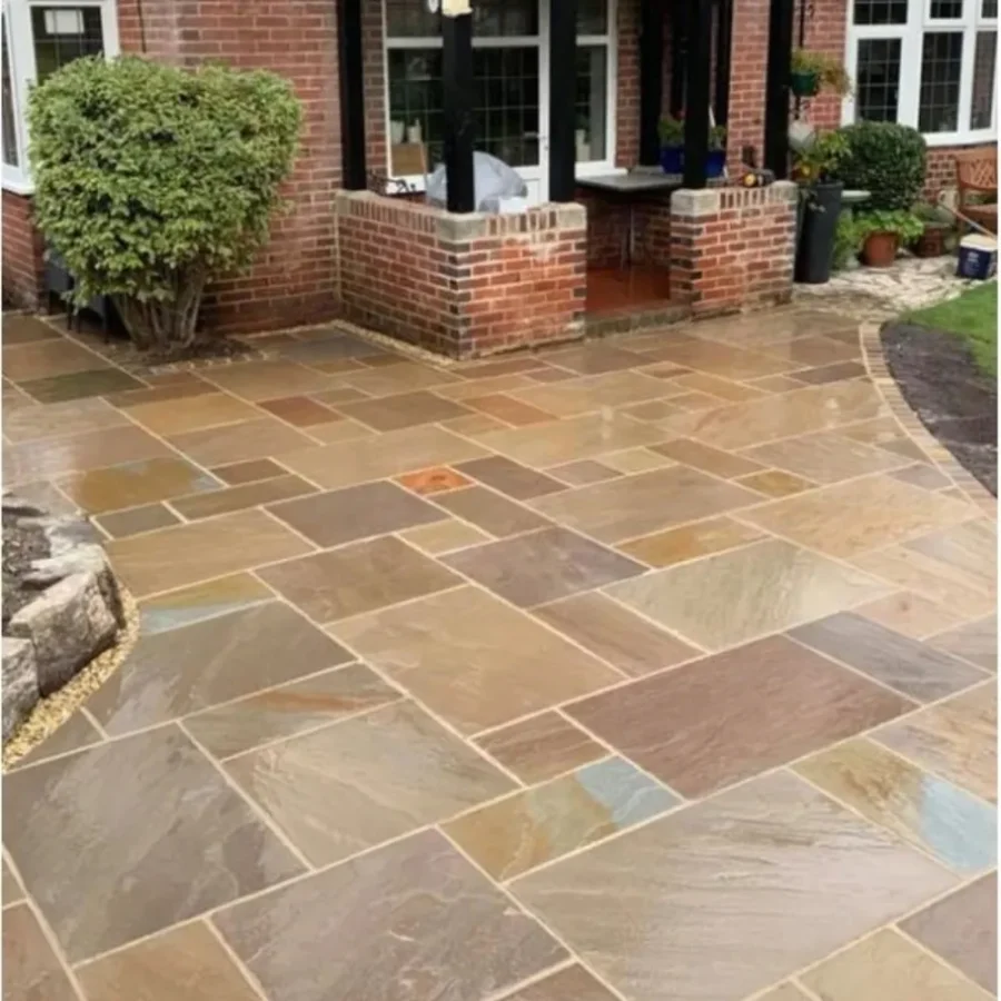 A freshly paved patio features Autumn Brown Indian Sandstone Paving Slabs in hand-cut, mixed sizes, laid out in geometrical shapes next to a brick house. The area is adorned with a small bush and potted plants, creating a tidy and inviting outdoor space.