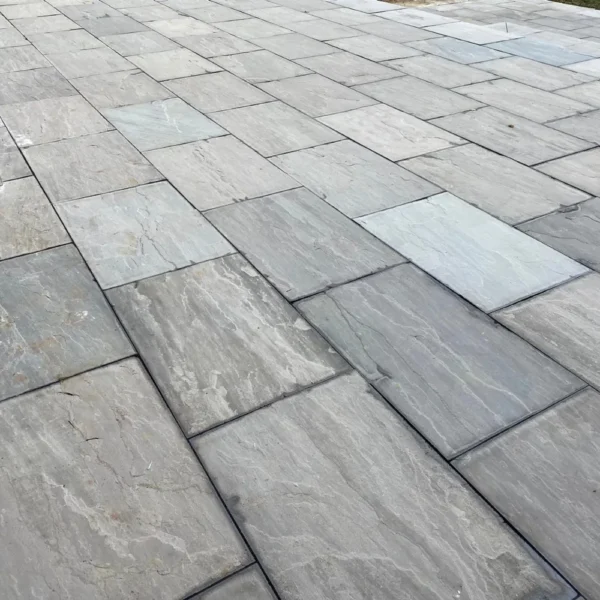 Detailed view of a patio constructed with Kandla Grey Sandstone Sawn Edge pavers, size 600mm x 900mm and 22mm thick. These rectangular stones, showcasing varying shades of gray, are arranged in a staggered pattern. The surface is smooth with visible natural textures and slight color variations.