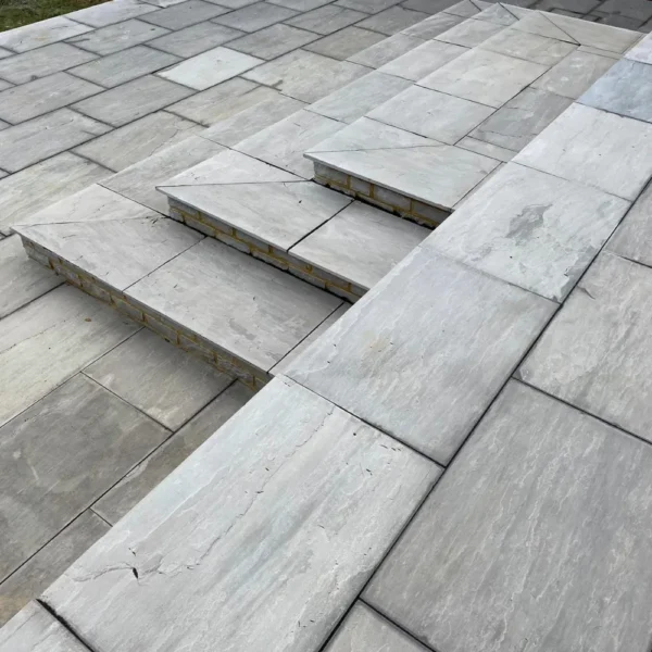 A set of three steps crafted from Kandla Grey Sandstone Sawn Edge tiles, measuring 22mm in thickness and arranged neatly into a rectangular pattern, leading up to a stone-tiled patio. The light gray tiles have a slightly textured surface, creating an inviting outdoor area.