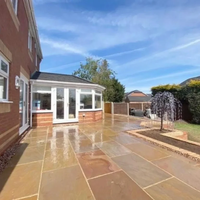 A modern brick house showcases an expansive patio made of Raj Blend Sandstone - 18mm Calibrated 600mm x 900mm slabs, complemented by a small landscaped area featuring a young tree. The clear, blue sky enhances the bright and inviting ambiance of this outdoor space.