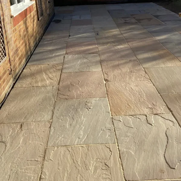 A sunlit patio featuring Raj Green sandstone paving, measuring 22mm in thickness and 600 x 900mm in size, displays an array of beige and light brown hues. The tiles have textured surfaces and are meticulously arranged in a rectangular pattern adjacent to a brick wall.