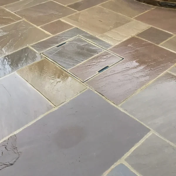 Raj Green - 22mm - Calibrated Natural Mixed Size paving slabs feature a concealed square drainage cover. The slabs come in various natural shades of tan, brown, and gray, with a smooth, glossy finish that reflects light.