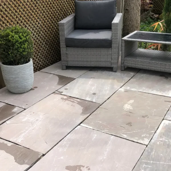 Outdoor seating area with a wicker chair and gray cushions on an Autumn Brown Sandstone 22mm - Calibrated 600mm x 900mm patio. A matching gray side table and a potted plant are nearby. The setting is bordered by a wooden lattice fence.