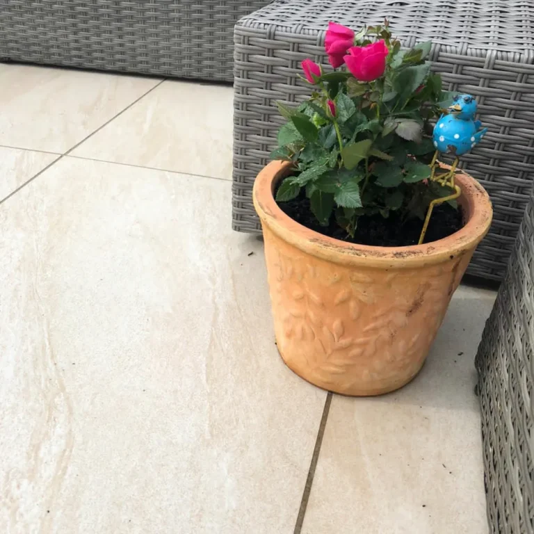 A terracotta pot with pink roses and a small blue decorative frog is placed on County Crema Outdoor Porcelain Paving Tiles in a light tiled patio setting, nestled between woven gray outdoor furniture.