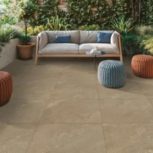 Contemporary outdoor patio with large Mocha Outdoor Porcelain Paving Tiles (600mm x 900mm), a light gray sofa with blue and gray pillows, a small round coffee table, two round wicker poufs, potted plants, and lush green foliage along a brick wall.
