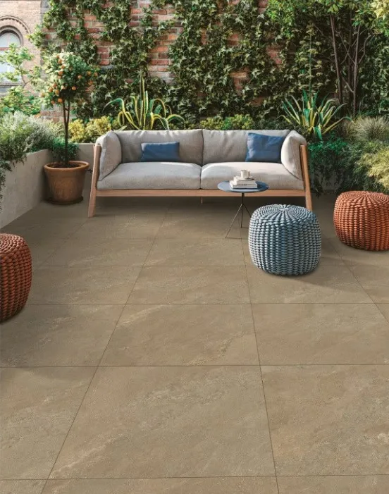 Contemporary outdoor patio with large Mocha Outdoor Porcelain Paving Tiles (600mm x 900mm), a light gray sofa with blue and gray pillows, a small round coffee table, two round wicker poufs, potted plants, and lush green foliage along a brick wall.